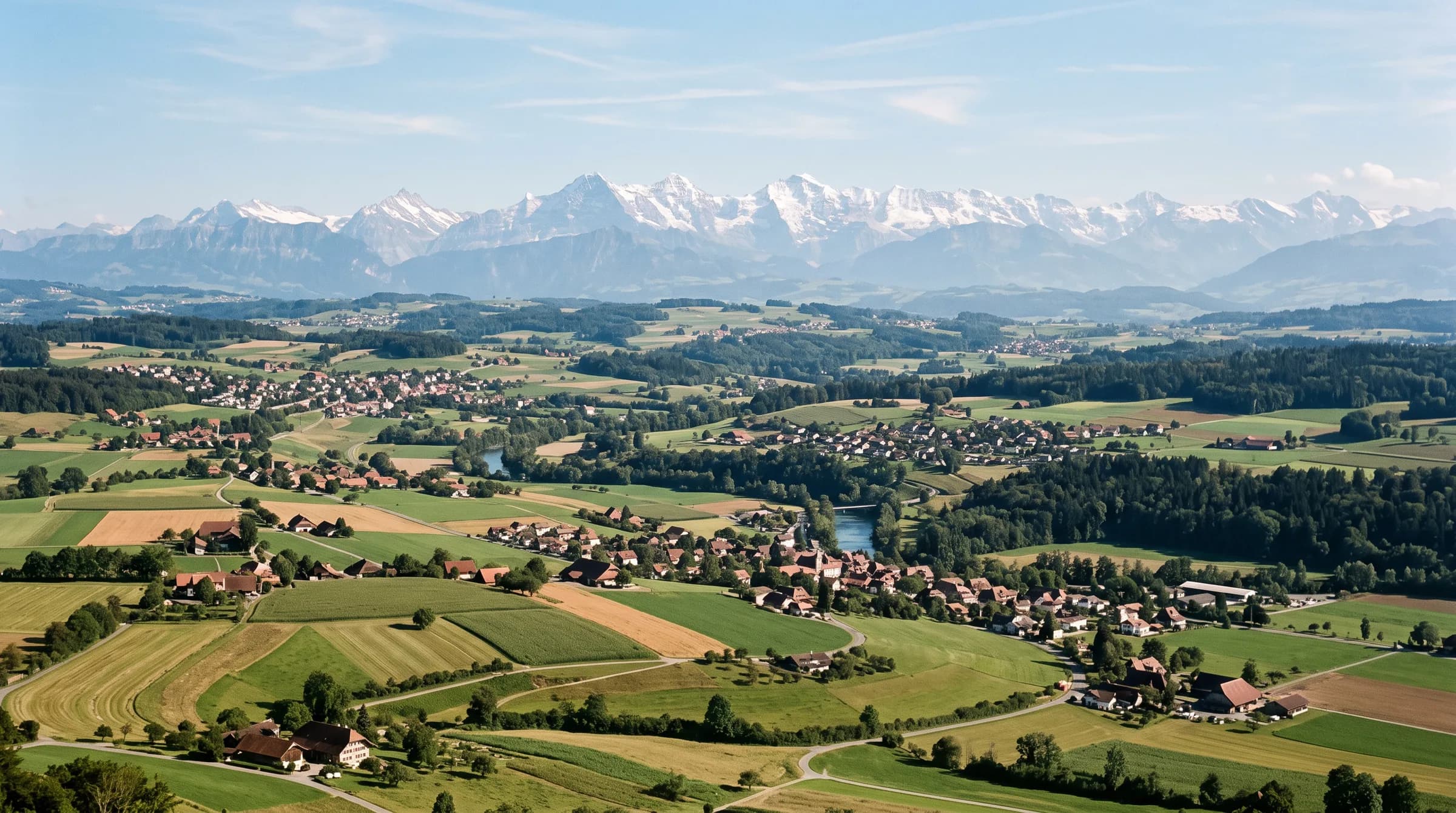 Weite Sicht über das Mittelland mit Feldern, Dörfern und bewaldeten Hügeln, am Horizont die verschneite Alpenkette.